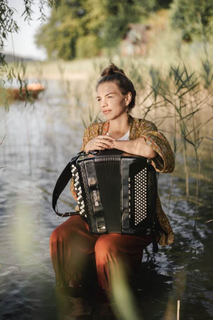 Bild der Musikerin maola mit Akkordeon am Ufer eines Sees im Wasser sitzend